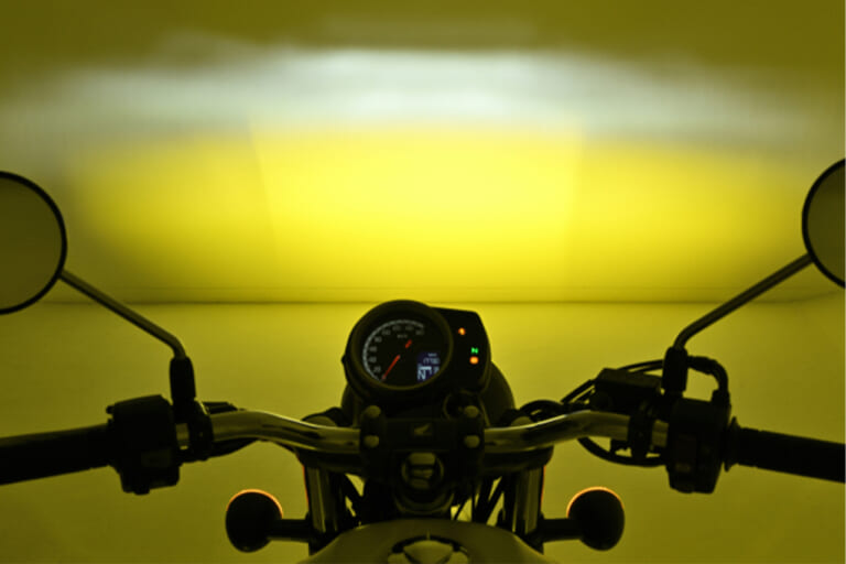 デイトナのLEDフォグランプユニット プレシャスレイ スモール イエローをバイクに装着し、白い壁に照射している写真｜「スイッチONで明らかに違いを感じる…」あらゆる車種に取付OK。コンパクトボディながら1700ルーメンで視界を確保する［デイトナ・LEDフォグランプユニット プレシャスレイ スモール］