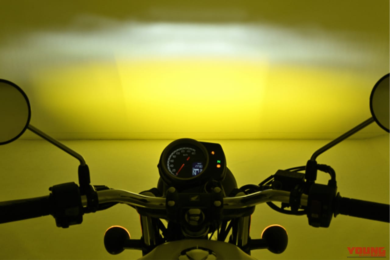 デイトナのLEDフォグランプユニット プレシャスレイ スモール イエローをバイクに装着し、白い壁に照射している写真｜「スイッチONで明らかに違いを感じる…」あらゆる車種に取付OK。コンパクトボディながら1700ルーメンで視界を確保する［デイトナ・LEDフォグランプユニット プレシャスレイ スモール］
